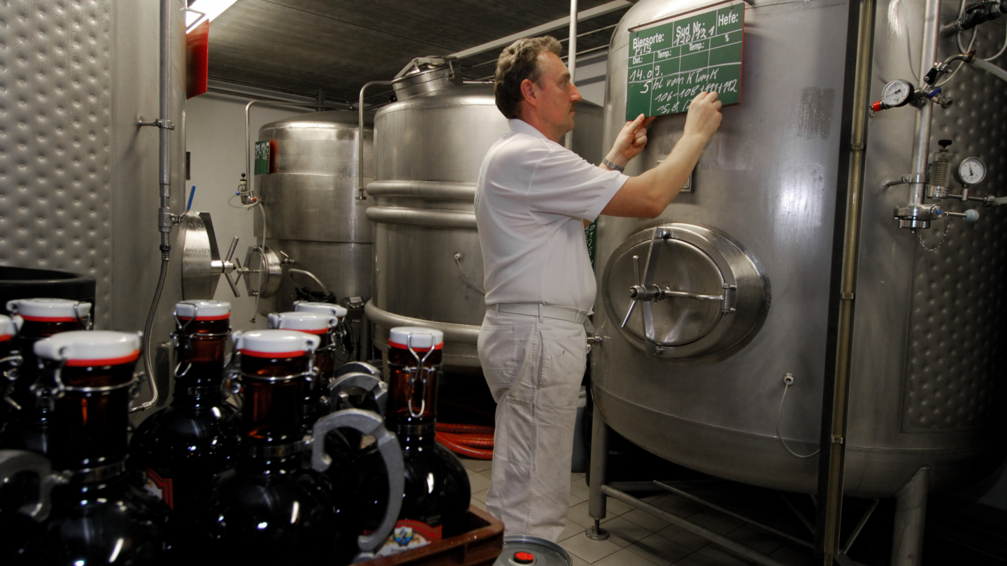 © Braugasthof Papiermühle, Foto: Rüdiger Widera Brauer steht vor Brauereikesseln mit Bierflaschen im Vordergrund © Braugasthof Papiermühle, Foto: Rüdiger Widera