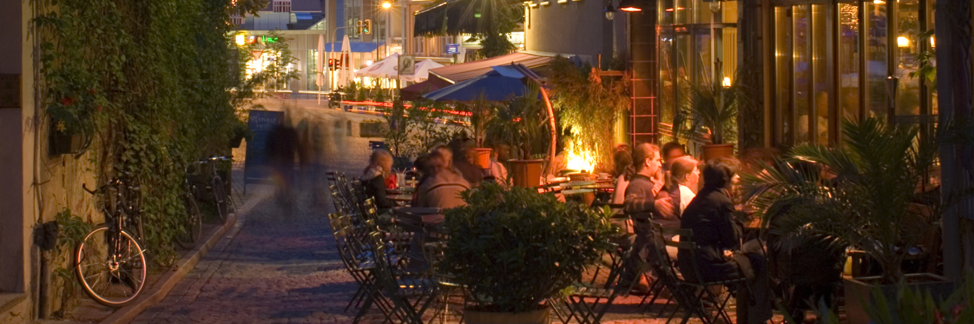 © JenaKultur, Foto: Toma Babovic Kulinarische Stadtführung: Die Wagnergasse in Jena bei Abendstimmung - Restaurantbesucher sitzen draussen und im Hintergrund leuchtet dies Fassade des JenTowers. © JenaKultur, Foto: Toma Babovic