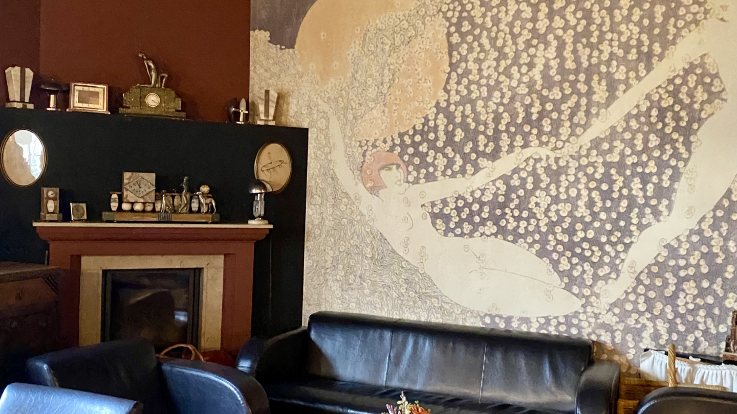 Das Kaminzimmer im Restaurant Bauersfeld des Planetariums Jena mit schwarzen Ledersofas, einem Holztisch und einer Wandmalerei mit zwei schwebenden Figuren ©JenaKultur_Foto: Jenny Marx